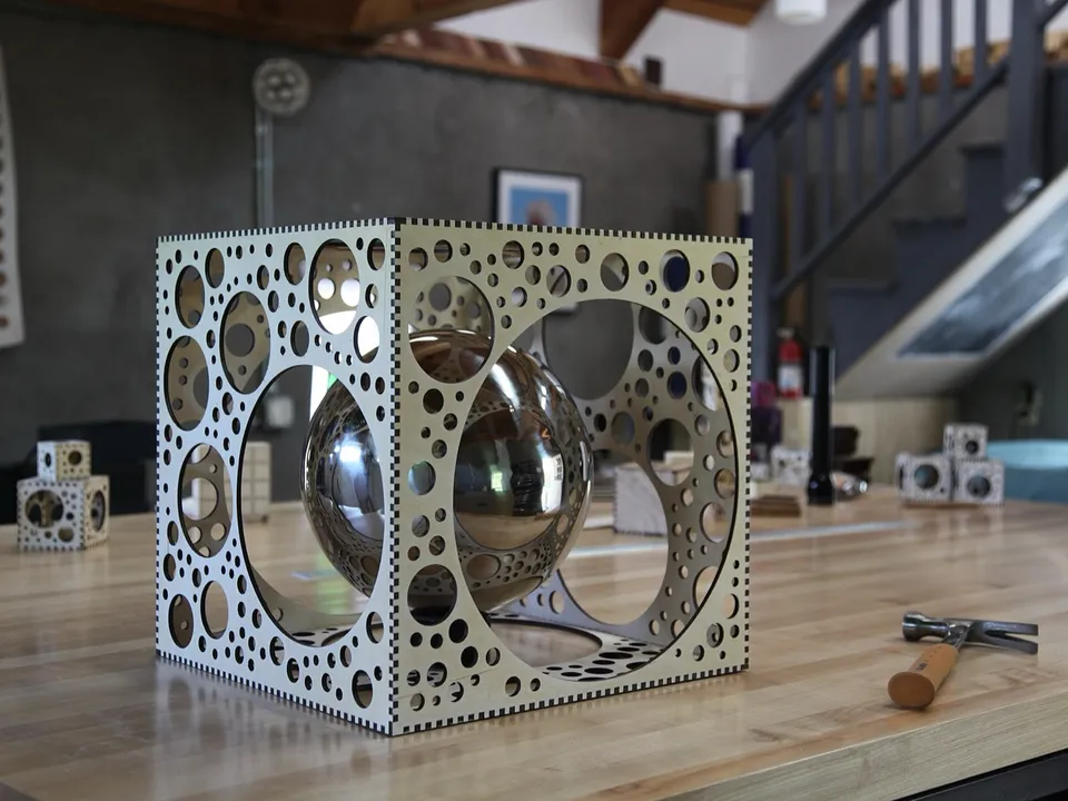 A large cube laser cut from birch plywood, its finger-jointed panels pierced with circular holes of many sizes, holding a mirrored sphere on a wooden workbench.