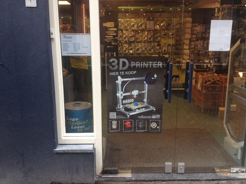 A Dutch-language poster reading '3D printer hier te koop' (3D printer for sale here) on the glass door of an electronics shop, showing a Velleman K8200 kit printer, with racks of electronic parts inside.