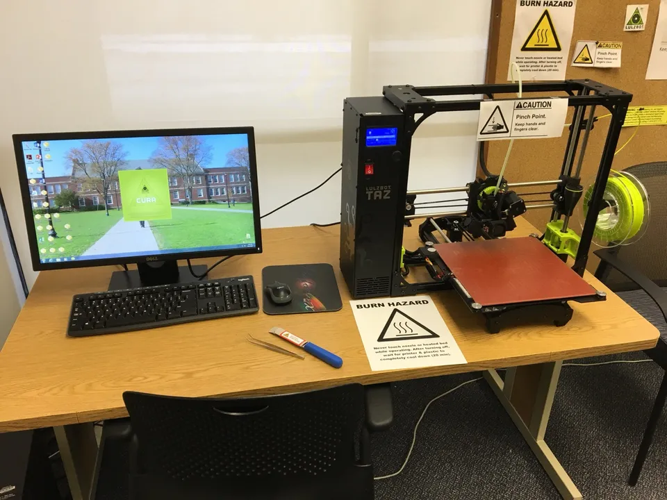 A LulzBot TAZ 6 3D printer on an office desk beside a monitor, keyboard and mouse, with a filament spool mounted on its side.