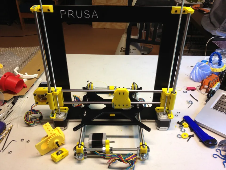 A partly built Prusa i3-style printer kit on a workbench: a black frame marked PRUSA, yellow printed parts, smooth and threaded rods and stepper motors, with loose nuts, washers and Allen keys around it.