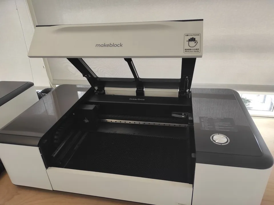 A Makeblock desktop laser engraver and cutter with its hinged lid raised over the work bed, on a shelf beside a window.