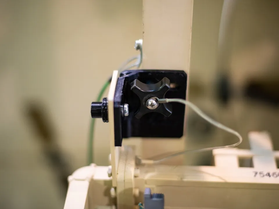 A black 3D-printed nylon mounting bracket with a cross-shaped knob, bolted to the beige frame of an antenna system, with a thin grey wire running past it.