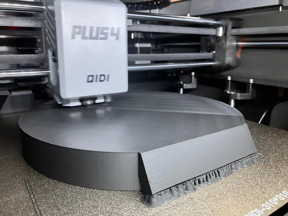A gray toolhead marked PLUS4 and QIDI printing a wide, dark gray disc-shaped part with a sloped edge on a textured build plate printed QIDI-310*310, inside the printer.