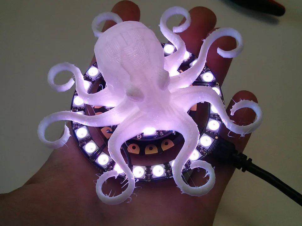A white 3D-printed octopus with curled tentacles sitting on a ring of lit LEDs in an open palm, with pliers on the table behind.