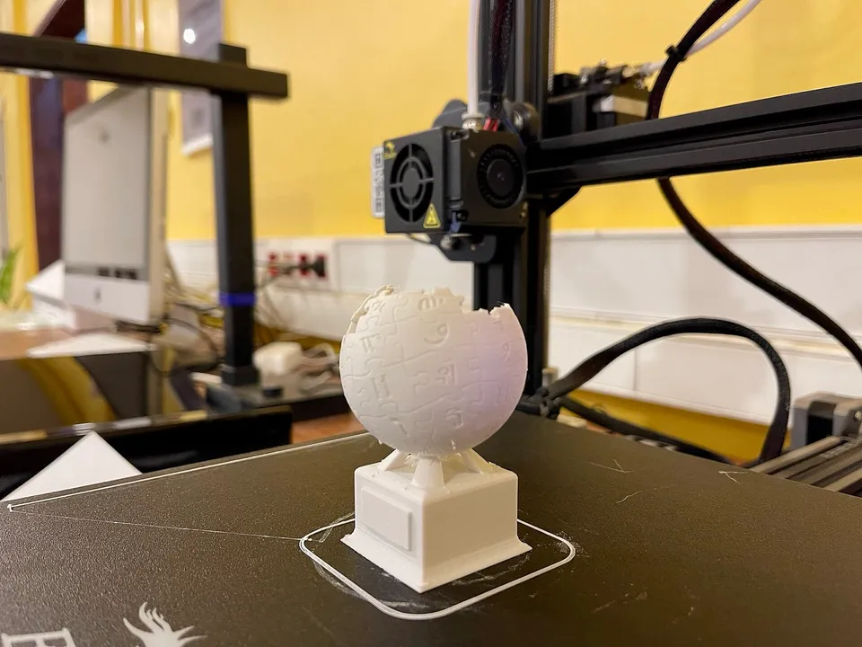 A white 3D-printed Wikipedia globe trophy on the black bed of an open-frame printer, with the toolhead and gantry above it.
