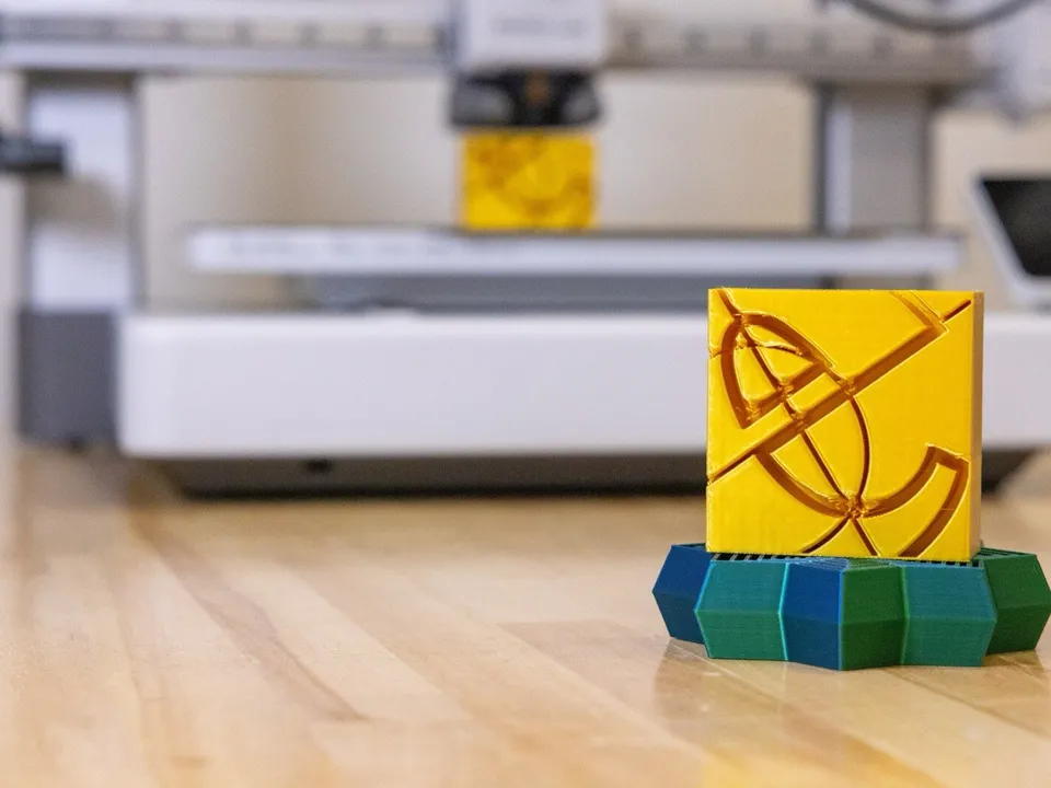 A yellow 3D-printed block on a blue and green printed base on a wooden table, with a white Bambu Lab A1 bedslinger out of focus behind it and a matching yellow print on its bed.