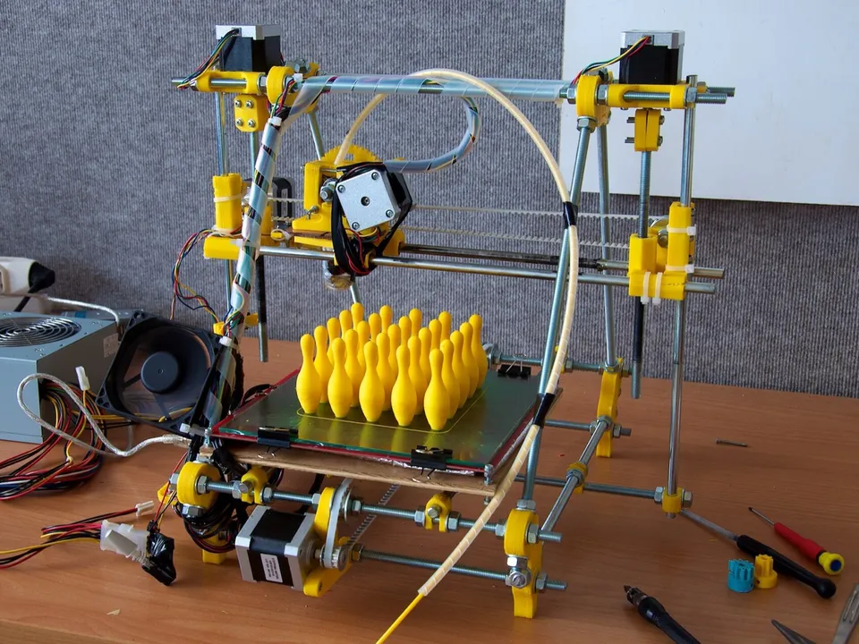 A home-built Prusa Mendel RepRap with yellow printed parts and a threaded-rod frame, a batch of small yellow prints on its bed and hand tools on the desk around it.