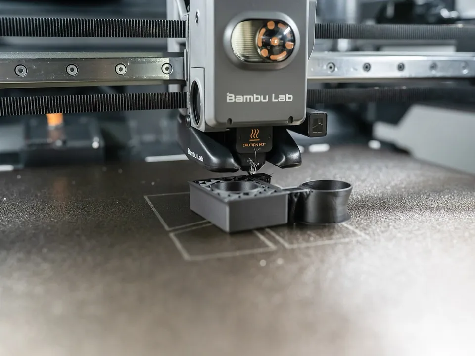 Close-up of a Bambu Lab toolhead laying down a dark grey part on a textured build plate, with belts and a linear rail behind it.