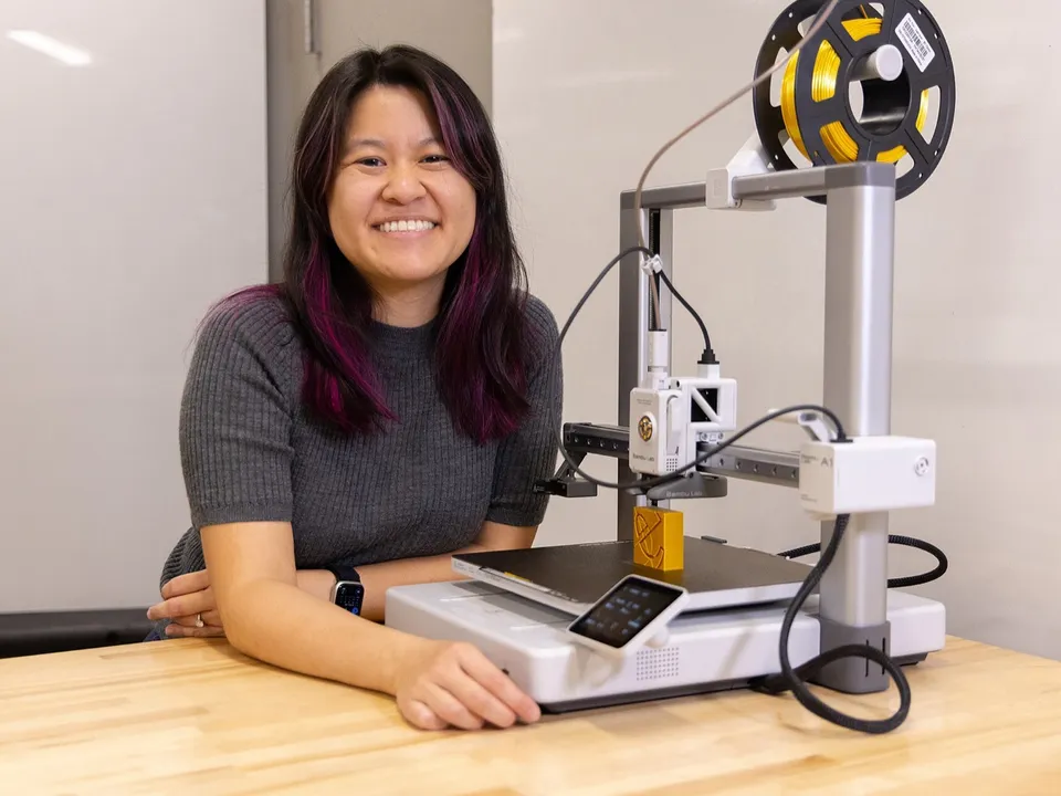 A smiling woman seated at a wooden workbench beside a white Bambu Lab A1 bed-slinger, with a yellow filament spool on top and a small yellow print on the bed.
