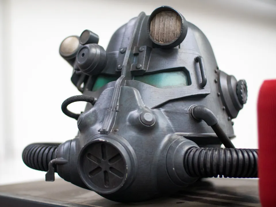 A painted 3D-printed power armor helmet from the Fallout game series, with a round filter on the mouthpiece and ribbed hoses at each side, on a dark table.