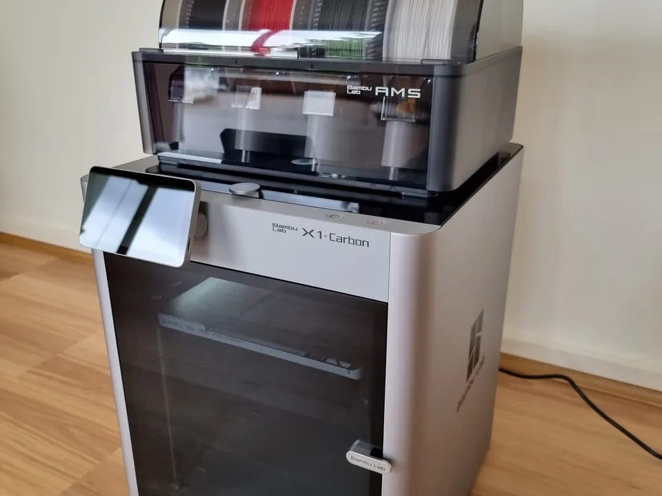 A Bambu Lab X1-Carbon enclosed printer with an AMS unit holding four filament spools on top, standing on a wooden floor in a home.