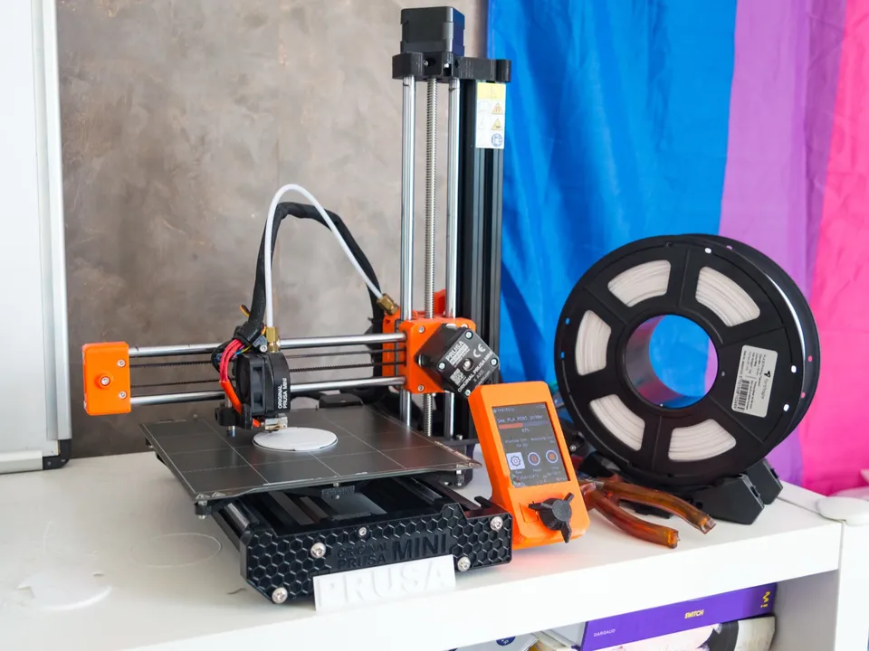 An orange and black Prusa Mini on a shelf, its X arm with the toolhead held out over the bed from a single vertical Z column at the back right.
