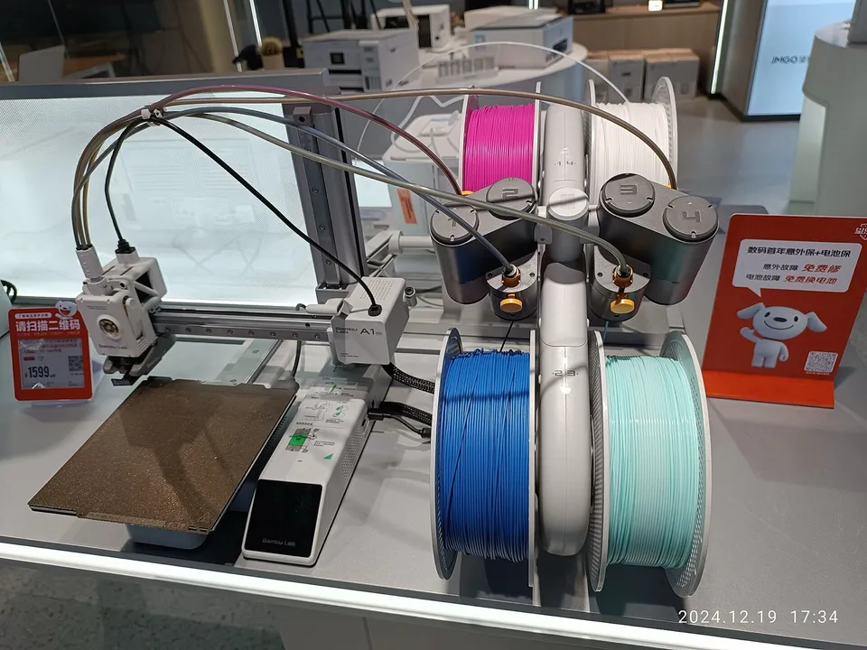 A Bambu Lab A1 mini on a store counter beside its AMS lite unit, which holds magenta, white, blue and mint filament spools, with a price tag on the left.