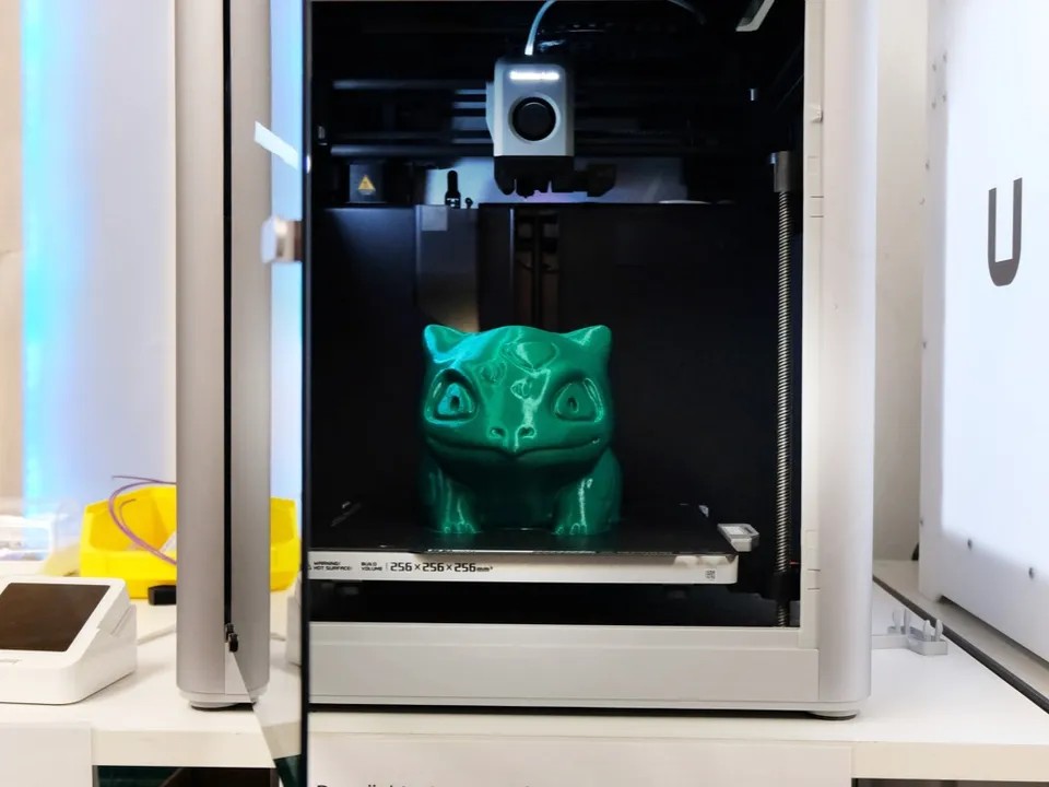 A Bambu Lab X1-Carbon with its door open and an AMS unit on top, the CoreXY toolhead parked above a green print on the bed.