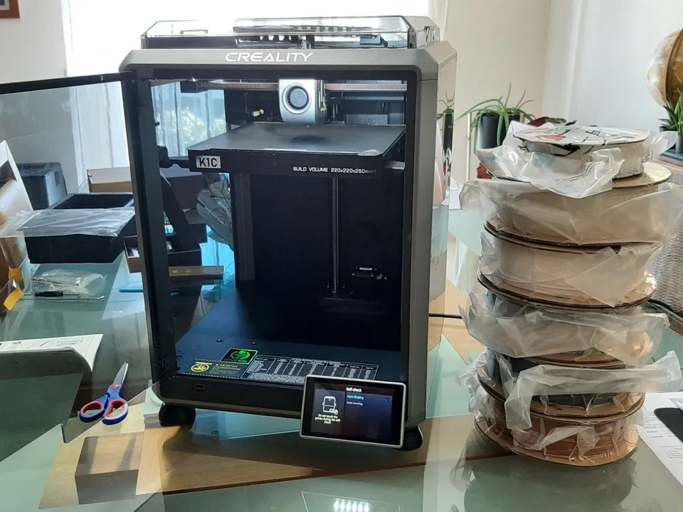 An enclosed Creality K1C CoreXY printer on a glass table with its door open, the toolhead on the gantry above the bed, beside a stack of wrapped filament spools.