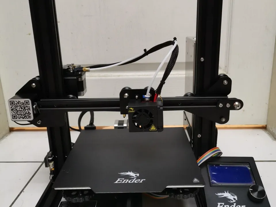 A fully assembled Creality Ender 3 on a white tiled floor, seen from the front: the black aluminium frame, the hotend on the X gantry, the Ender-branded bed and the control box with its blue LCD screen at right.
