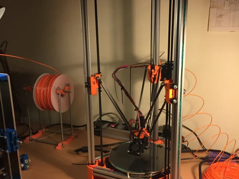 A tall self-built Kossel delta printer on a workbench, with three arms holding the hotend over a round glass bed inside an aluminum frame with orange printed corners.