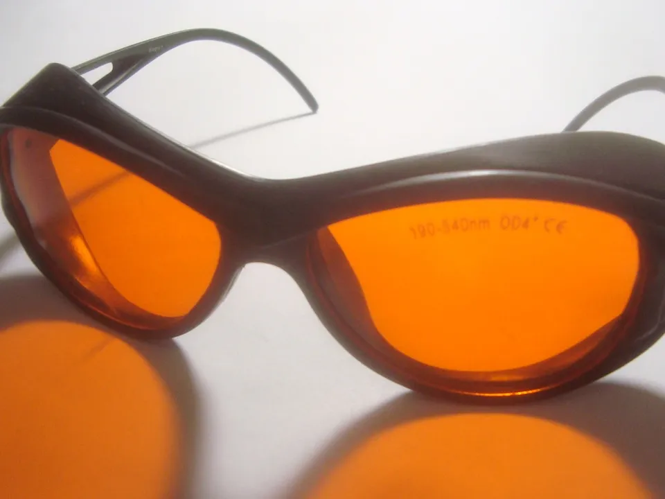 A pair of orange-lensed laser safety goggles marked 190-540nm OD4+ CE, resting on a white surface.