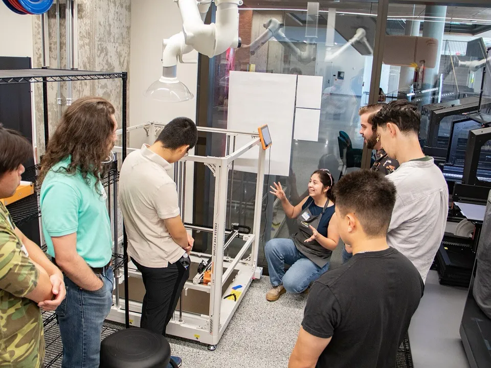 A woman crouches beside a large white open-frame Elegoo printer with an orange touchscreen and toolhead, explaining it to a group of students in a university lab, with enclosed printers along the glass wall.