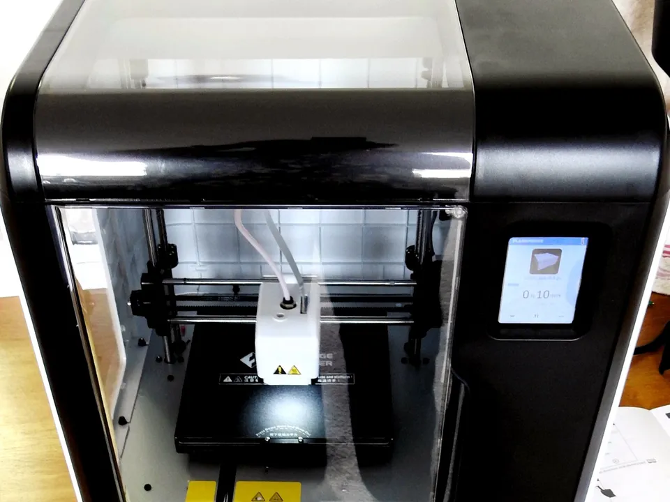 A Flashforge Adventurer 3 on a wooden desk, its clear front door and top window showing the print head over the build plate, with the touchscreen on the right.