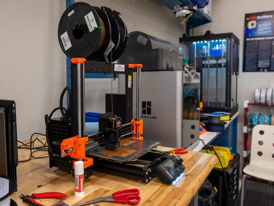 An open-frame Original Prusa MK4 on a workbench in front of an enclosed Bambu Lab printer and a tall enclosed printer