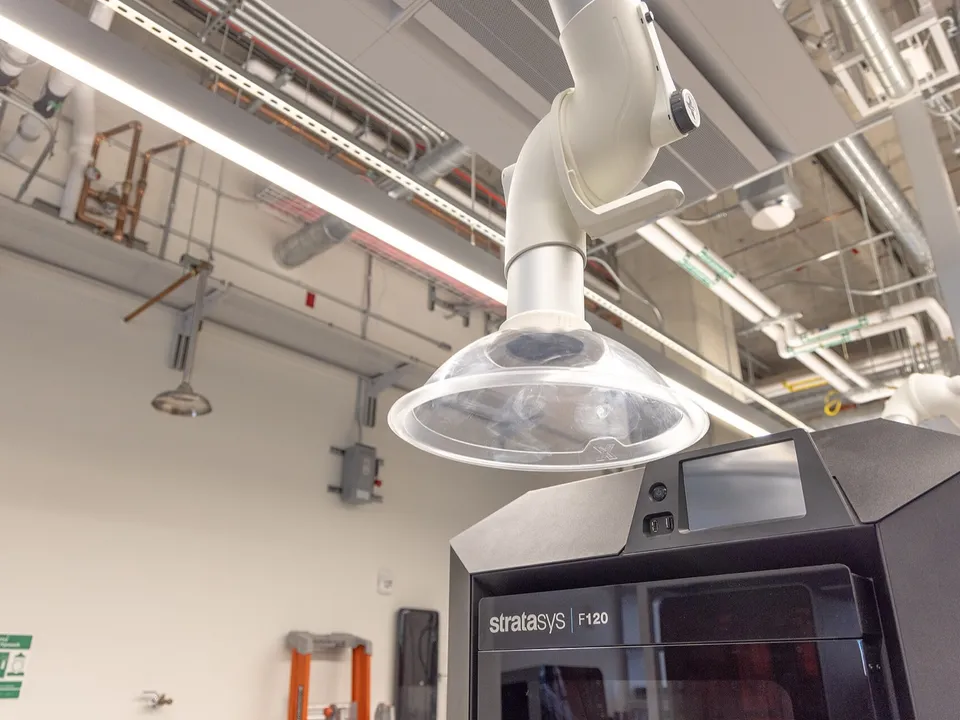 A ceiling-mounted fume extraction hood positioned over an enclosed Stratasys F120 3D printer in a university lab.