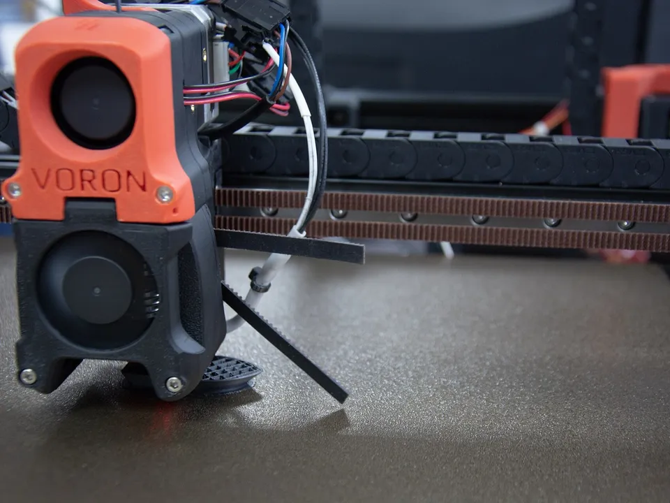 The orange Voron-branded toolhead of a Voron 3D printer on its gantry, with the cable chain and belt behind it and a textured build plate below.