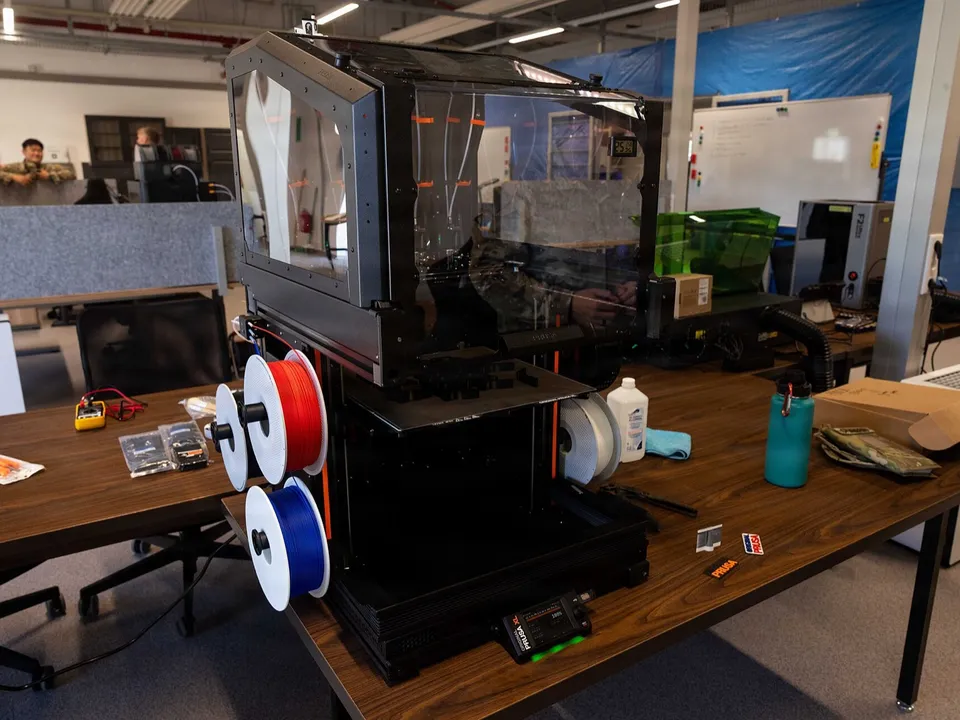 A large enclosed 3D printer on a workbench in an open-plan workshop, with red and blue filament spools hung on its side.