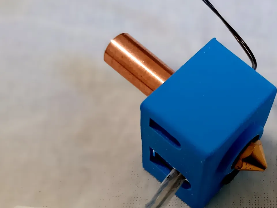 A loose 3D printer hotend on a white surface: a copper heat break, a heater block in a blue silicone sock with a brass nozzle, and the heater and sensor wires ending in white connectors.