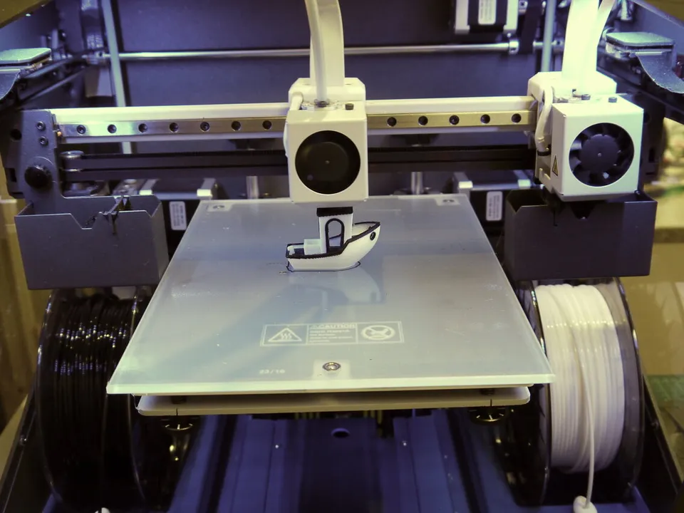 Front view of a BCN3D Sigma IDEX printer: one toolhead prints a black-and-white boat while the second sits parked at the right end of the shared X rail.