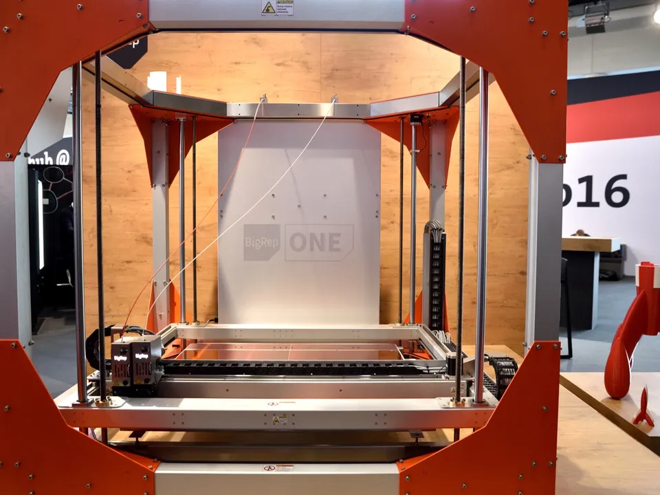 A BigRep ONE large-format 3D printer on a trade-show stand, its square orange-and-aluminum frame surrounding a wide print bed and long guide rails.
