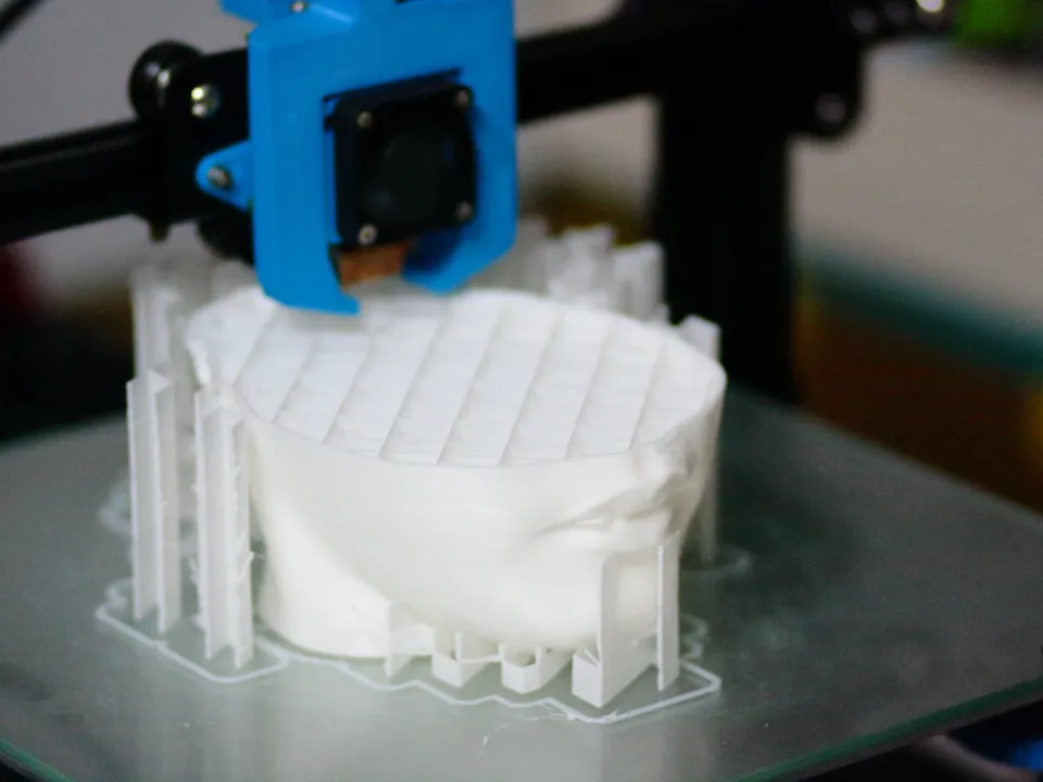 A life-size head half printed in white filament on the glass bed of a Creality CR-10, under a blue printed fan duct.