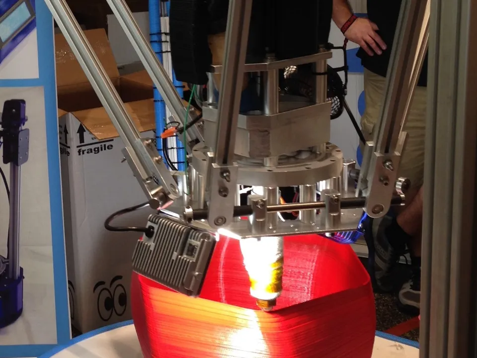 The effector of a large SeeMeCNC delta 3D printer laying down a tall red vase-shaped print on a round bed, its three arms hanging from aluminum towers.