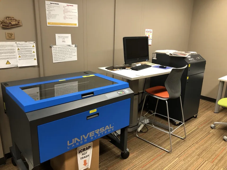 A Universal Laser Systems laser cutter in a university library makerspace, with a standalone fume extractor cabinet on the right and posted lists of approved and banned materials on the wall.