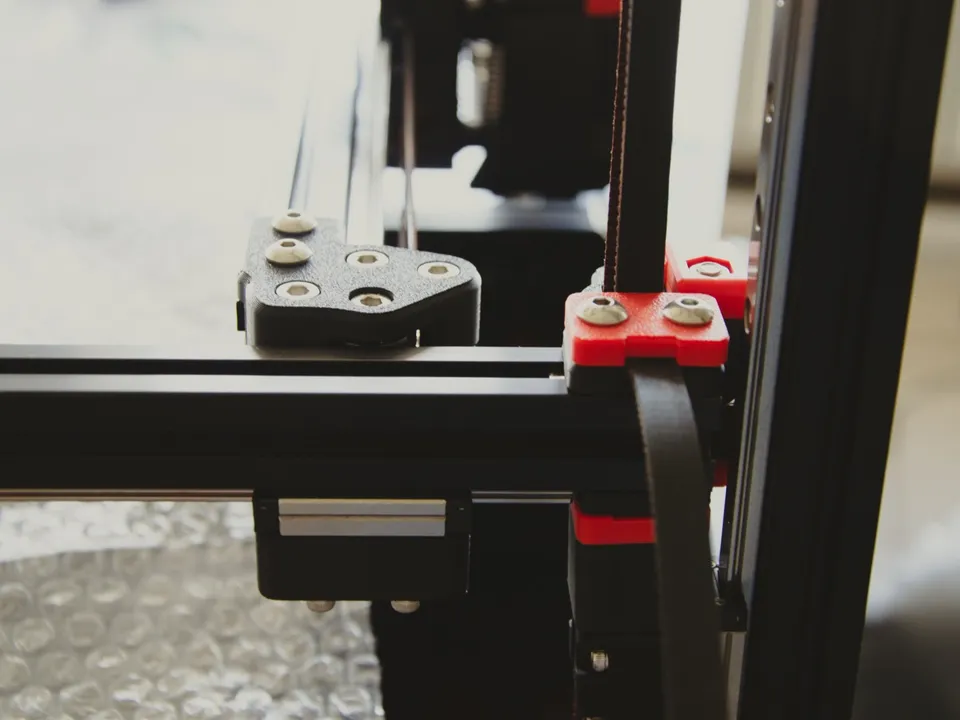 Close-up of a Voron 3D printer frame: a steel linear-rail carriage sits on its rail under a black aluminum extrusion, next to a belt and red printed parts.
