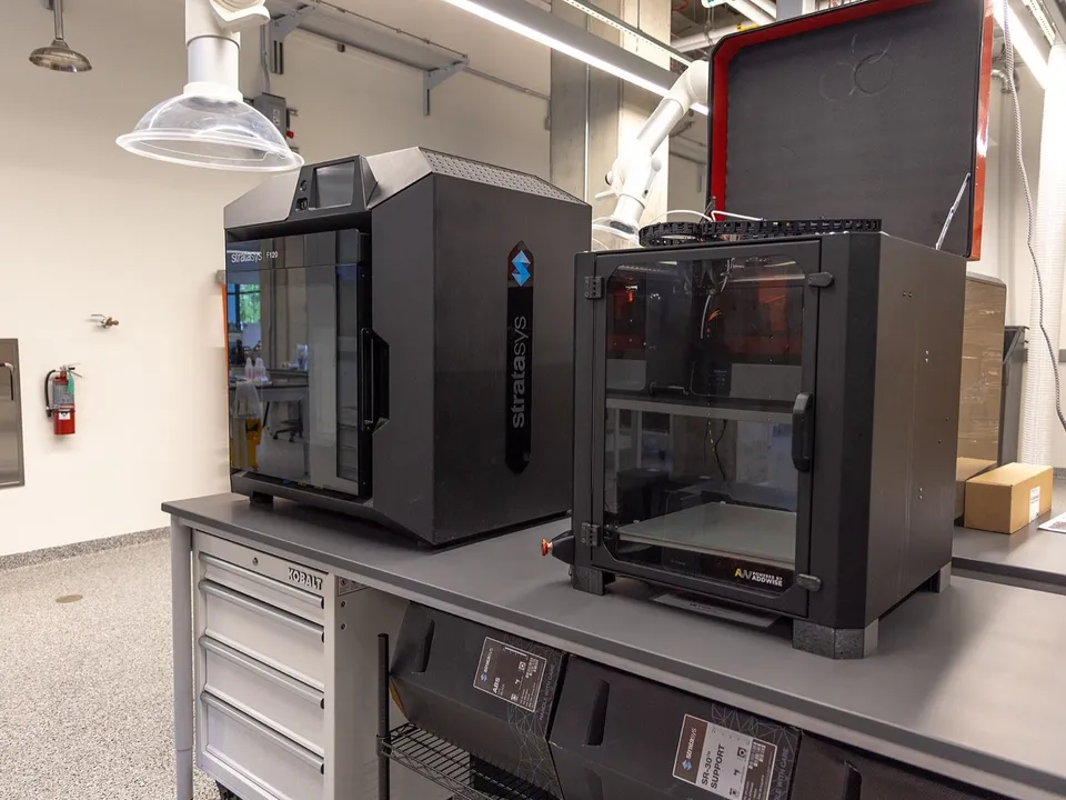 Two fully enclosed 3D printers, a Stratasys F120 and a second machine with its lid open, on a bench above boxed Stratasys ABS and support material.
