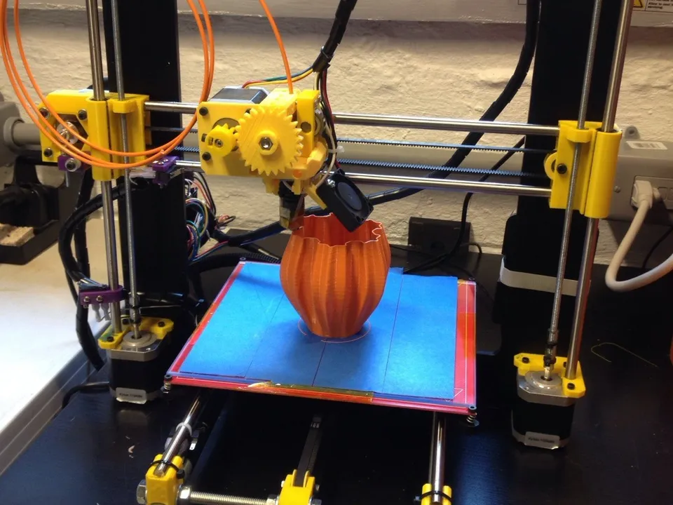 An open-frame Prusa i3 bedslinger printing an orange vase on a blue-taped bed that travels front to back on rods under the gantry.