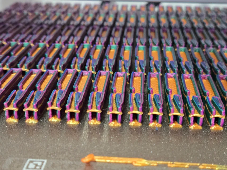 Rows of small 3D-printed whistles on a textured build plate, each printed in gold with purple, green and blue walls, with a gold line of filament on the plate in front.