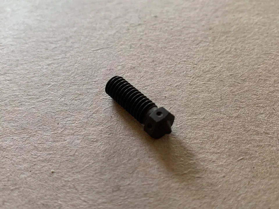 A single dark hardened steel 3D printer nozzle, with a threaded body and a hex-shaped tip, lying on a beige textured surface.