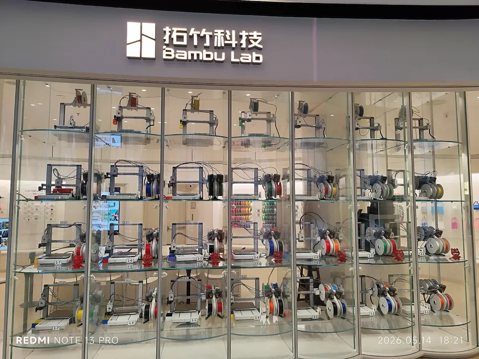 Rows of open-frame Bambu Lab bed-slinger printers, many with four-spool feeders beside them, on glass shelves in the window of a Bambu Lab shop in a Shenzhen mall.