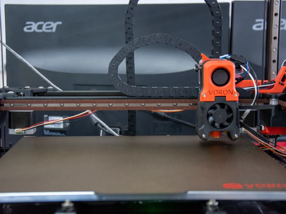 The empty textured build plate of a Voron 3D printer, with its orange toolhead parked above the plate.
