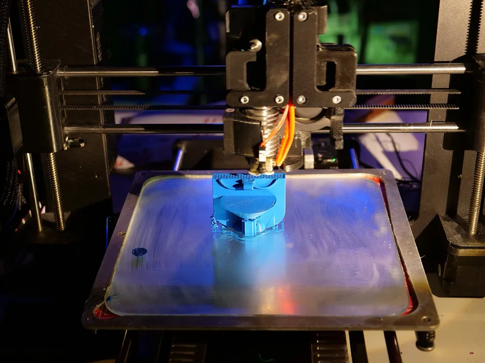 A desktop 3D printer printing a blue part on a heavily scratched build plate, with its belts, smooth rods and Z lead screws in view, at the 35th Chaos Communication Congress in Leipzig.