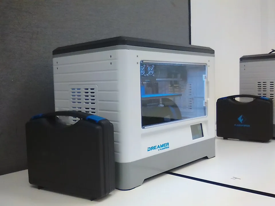 An enclosed Flashforge Dreamer printer on an office table beside its black carry case, with a second unit and case behind it