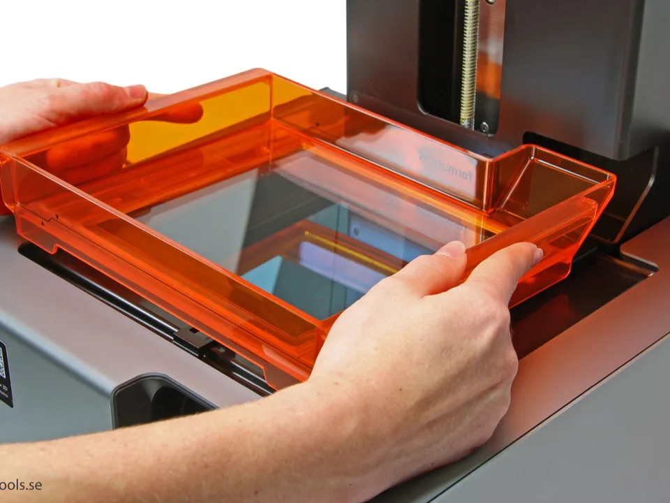 Two hands fitting the orange resin tank onto a Formlabs Form 2 stereolithography (SLA) 3D printer, below its build platform.