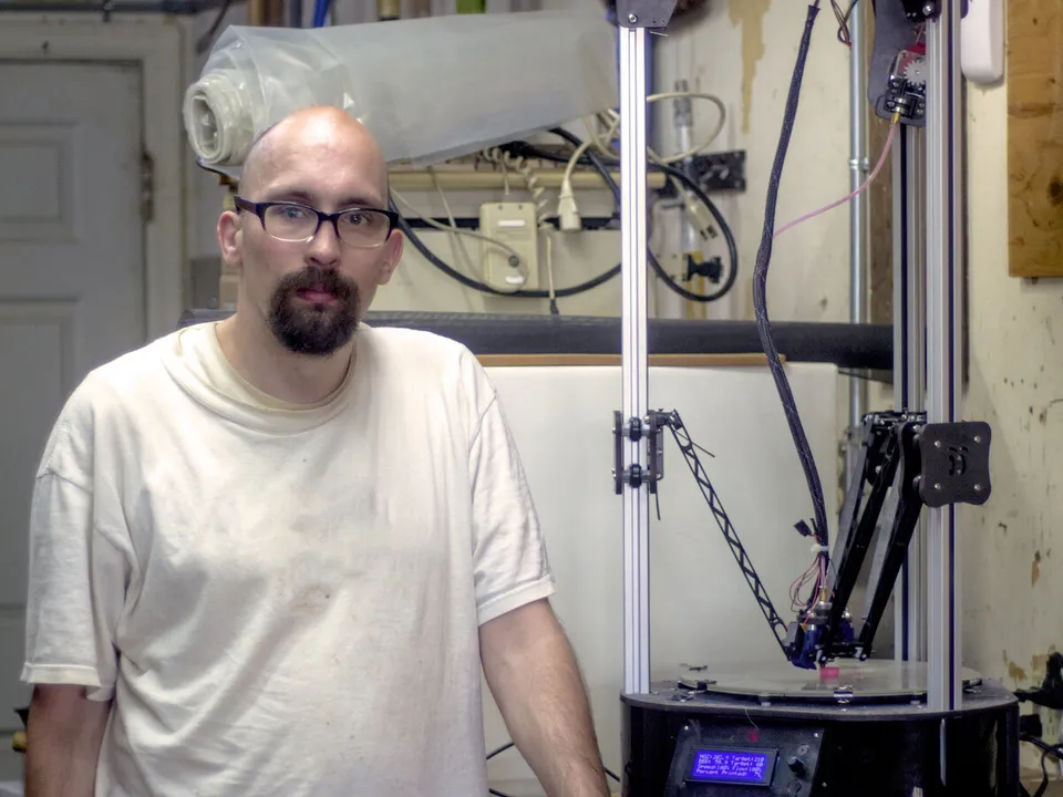 A man standing beside a SeeMeCNC Rostock Max V2 delta 3D printer on a workbench, its three arms hanging from carriages on three vertical towers above a round bed.