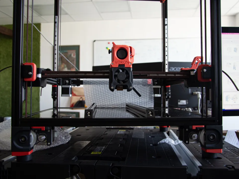 Front view of a Voron 3D printer's gantry: the red and black toolhead sits on its X beam, with belts running along the beam to pulley blocks at each end.