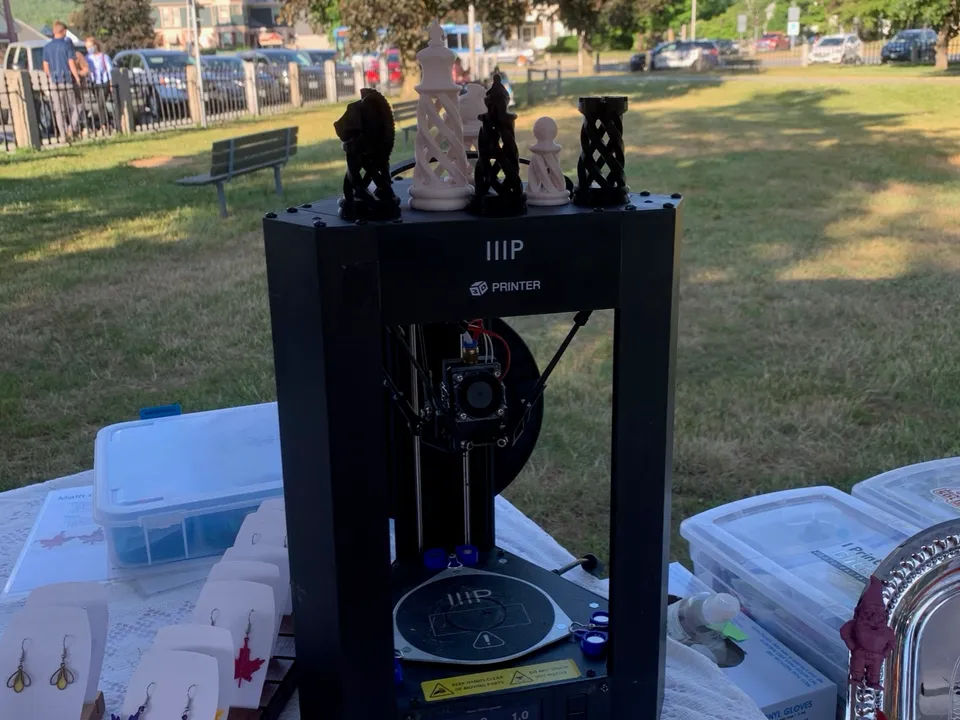 A compact black delta 3D printer on a vendor's table at an outdoor market on a town green, with printed chess pieces on top and printed earrings beside it.