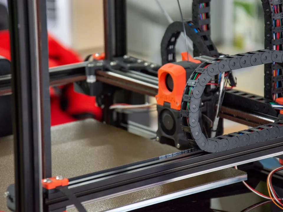 The orange toolhead of a Voron CoreXY printer on its X rail, with cable chains, above a textured build plate.
